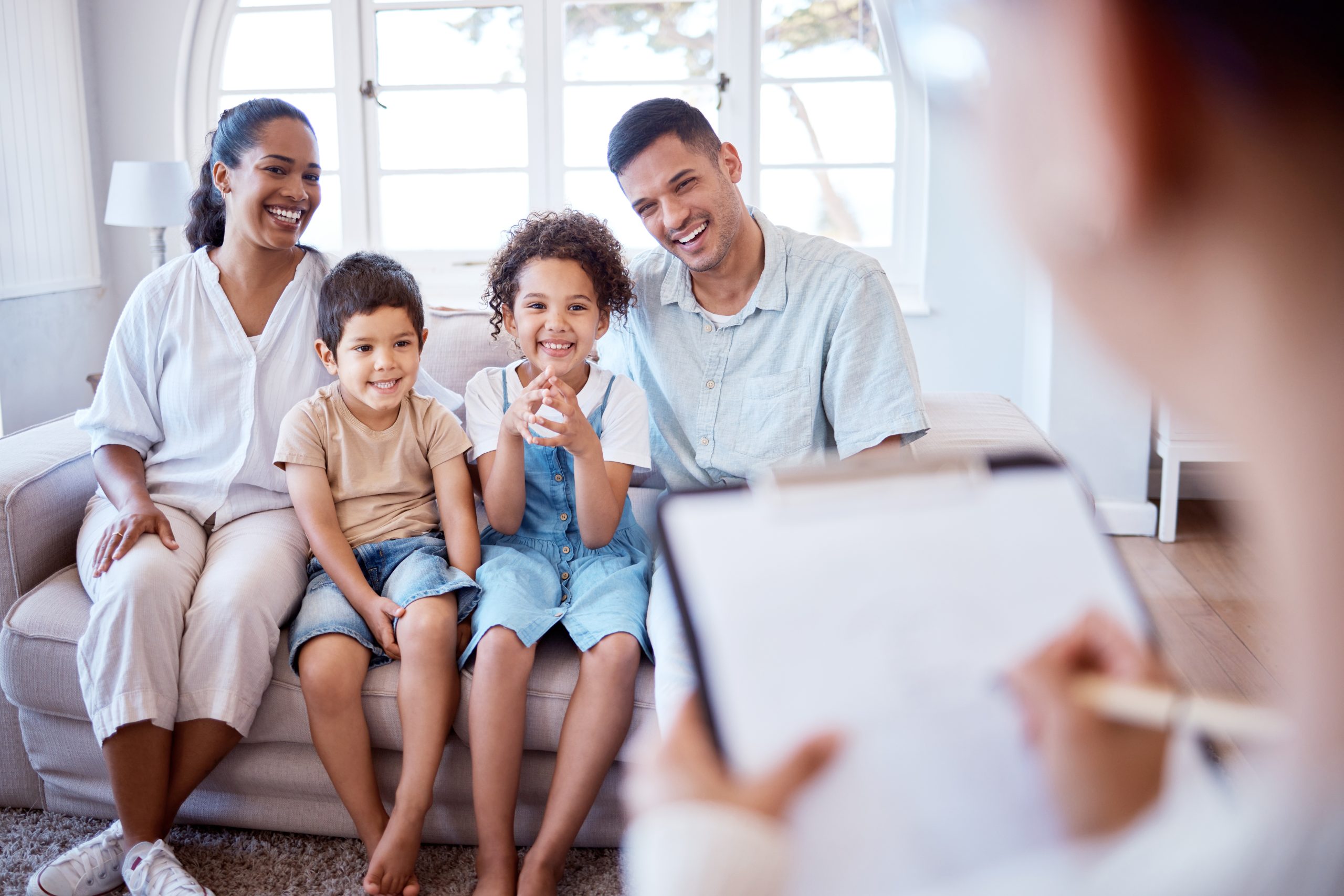 A family sits on a couch and a health care provider writes on a clipboard